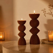 Two brown candle holders with lit candles on a wooden surface, with books and a vase in the background.
